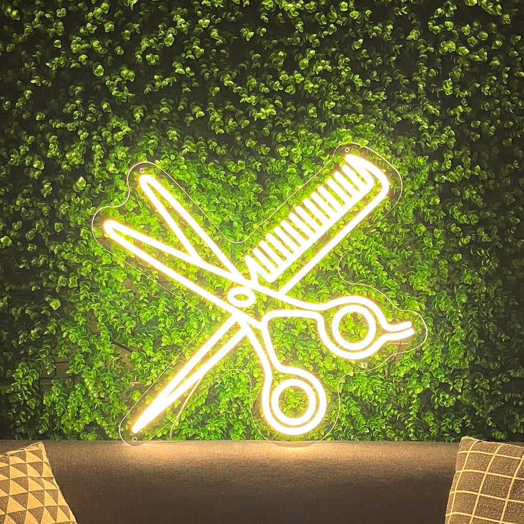 Neon sign of crossed scissors on a green leafy background above a sofa with patterned pillows.