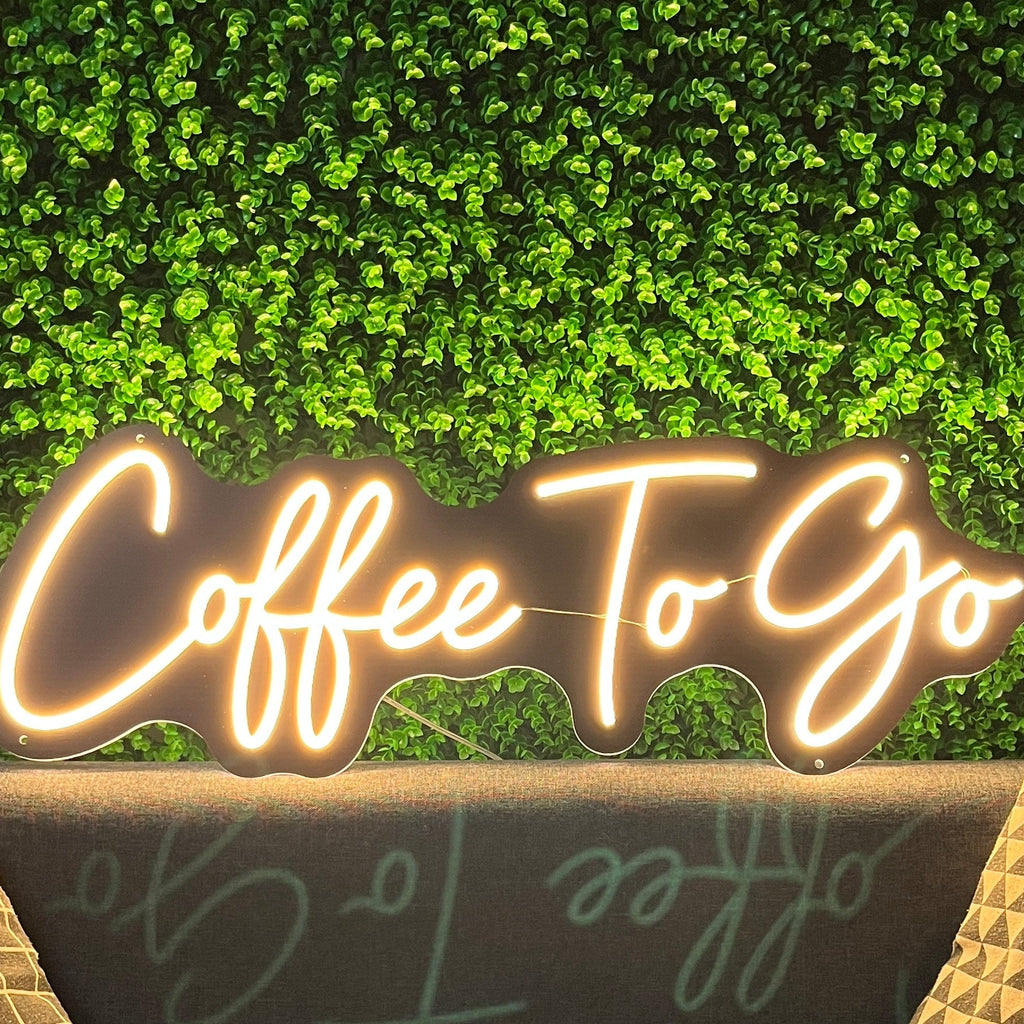 Neon sign reading 'Coffee To Go' above a couch with decorative pillows against a green wall.